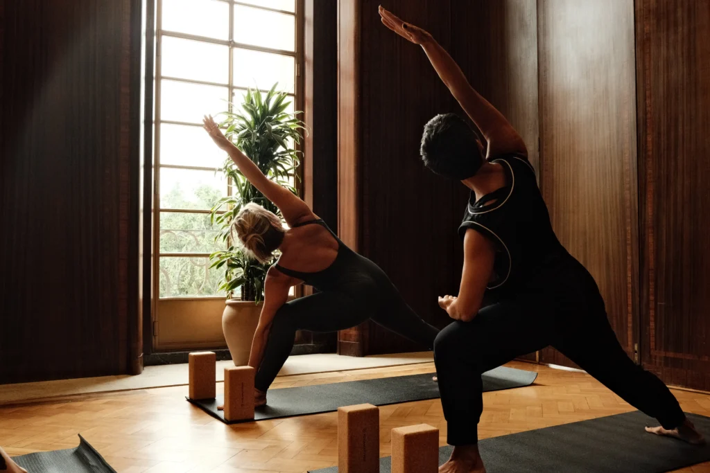 Two people practising yoga side by side on mats with cork blocks, beside a tall window